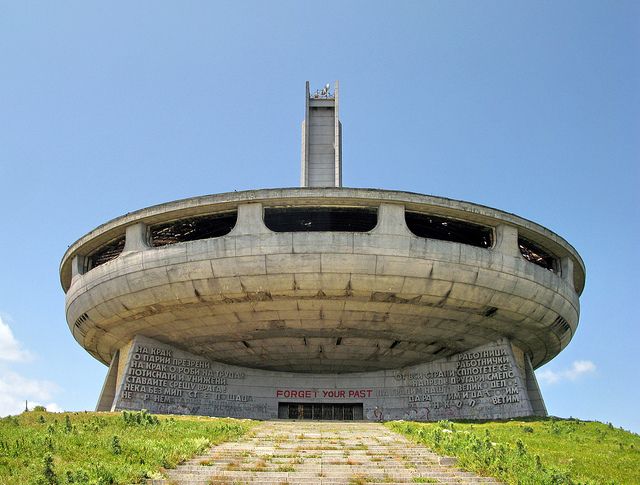 Low angle front view of the Buzludzha Monument entrance showing communist-era Cyrillic script and red FORGET YOUR PAST graffiti on the brutalist concrete.