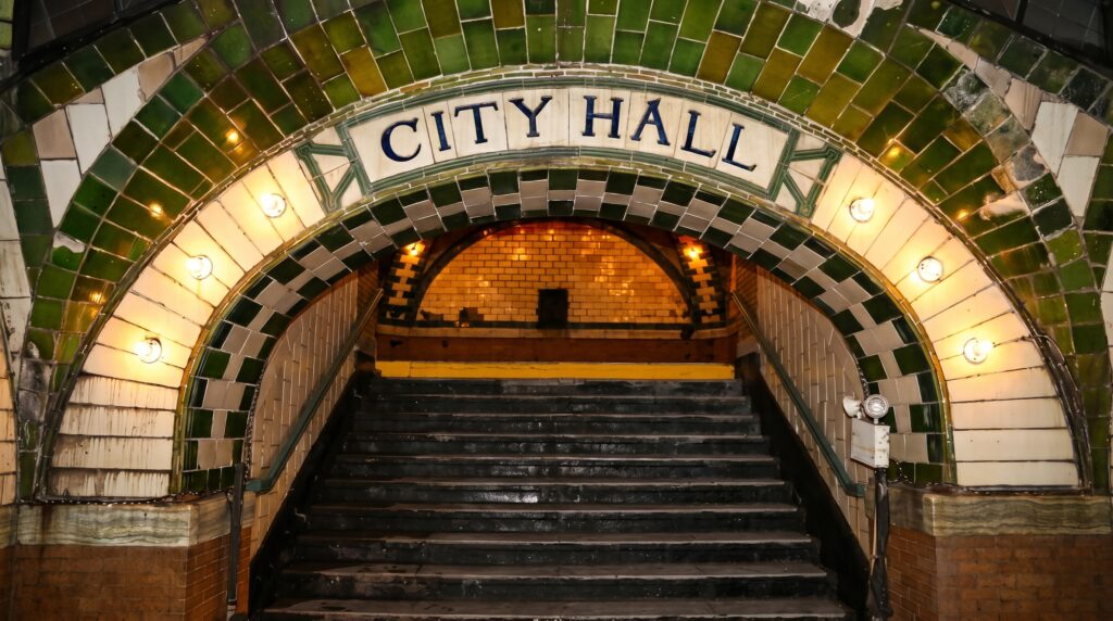 A wide, tiled archway featuring the words "CITY HALL" in blue lettering. The arch is bordered by green and white tiles and lined with round glowing lightbulbs, framing a dark staircase leading down into the station