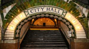 A wide, tiled archway featuring the words "CITY HALL" in blue lettering. The arch is bordered by green and white tiles and lined with round glowing lightbulbs, framing a dark staircase leading down into the station