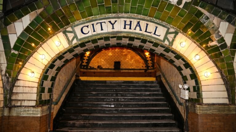 A wide, tiled archway featuring the words "CITY HALL" in blue lettering. The arch is bordered by green and white tiles and lined with round glowing lightbulbs, framing a dark staircase leading down into the station