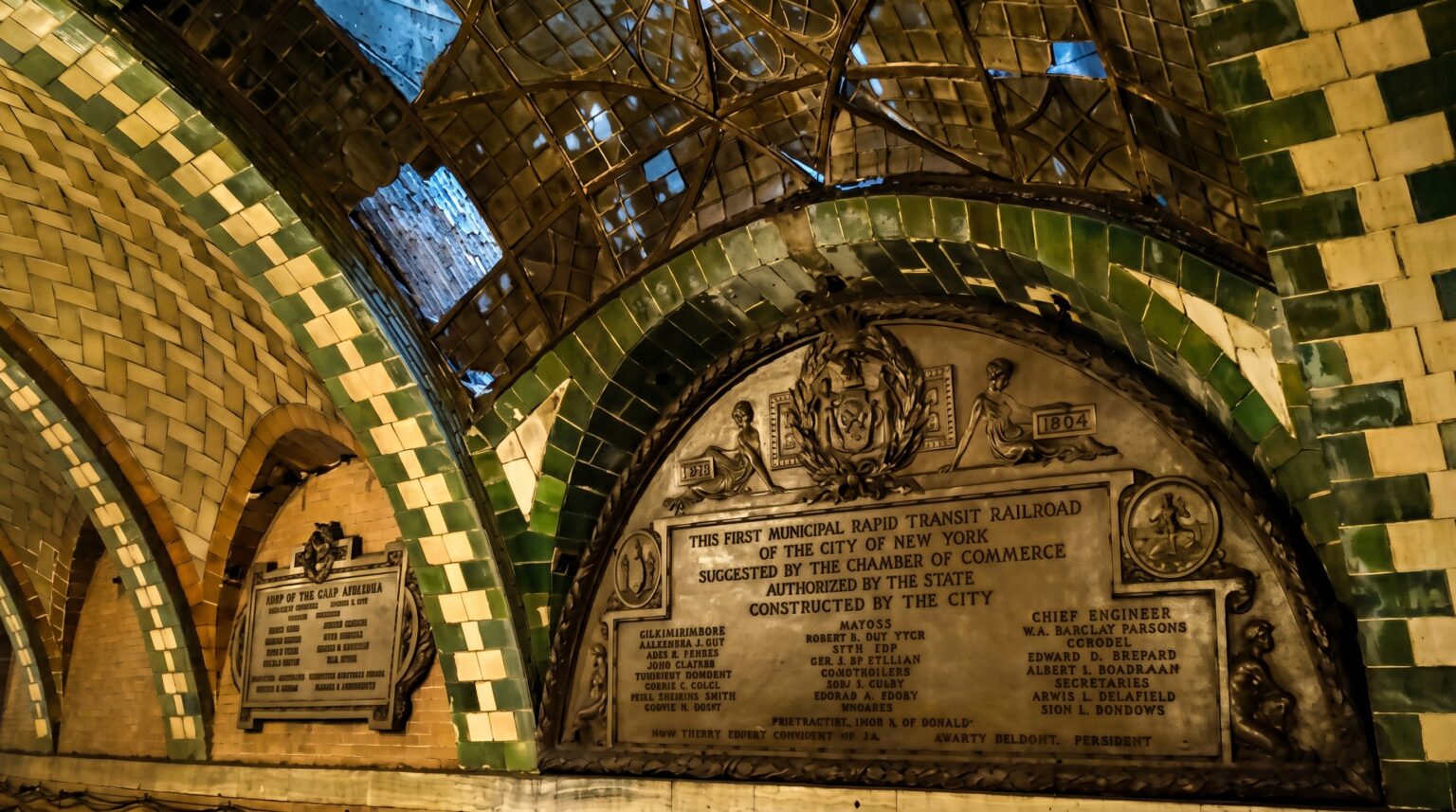 An ornate bronze plaque embedded in a green and cream tiled wall commemorating the "First Municipal Rapid Transit Railroad of the City of New York," situated beneath a damaged glass skylight