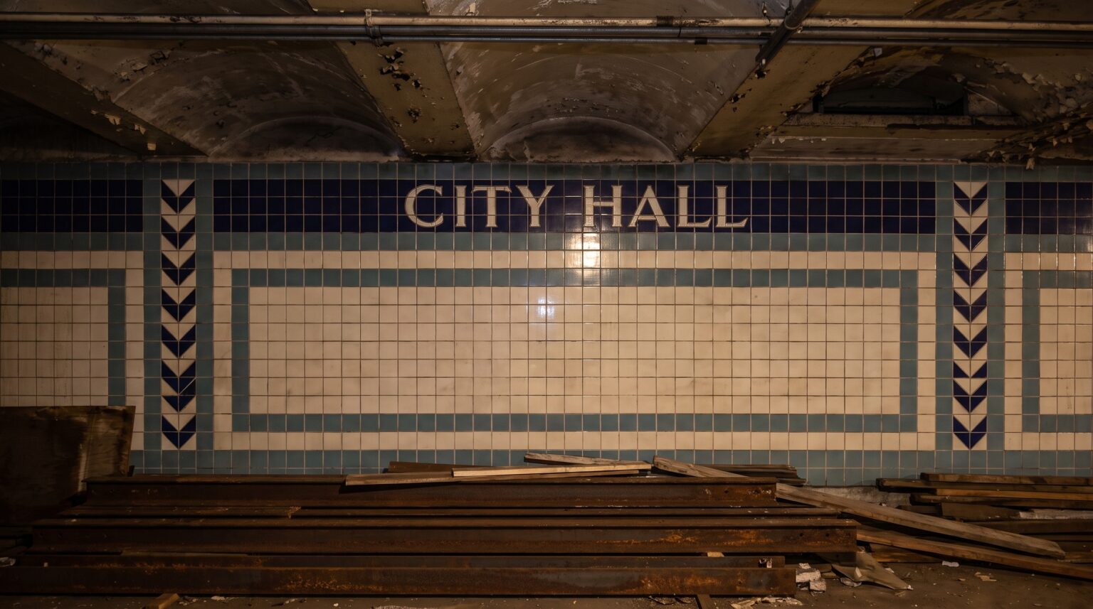 A tiled subway wall featuring the name "CITY HALL" in blue letters across the top, bordered by blue geometric and chevron patterns. A pile of rusted steel beams rests on the floor in the foreground