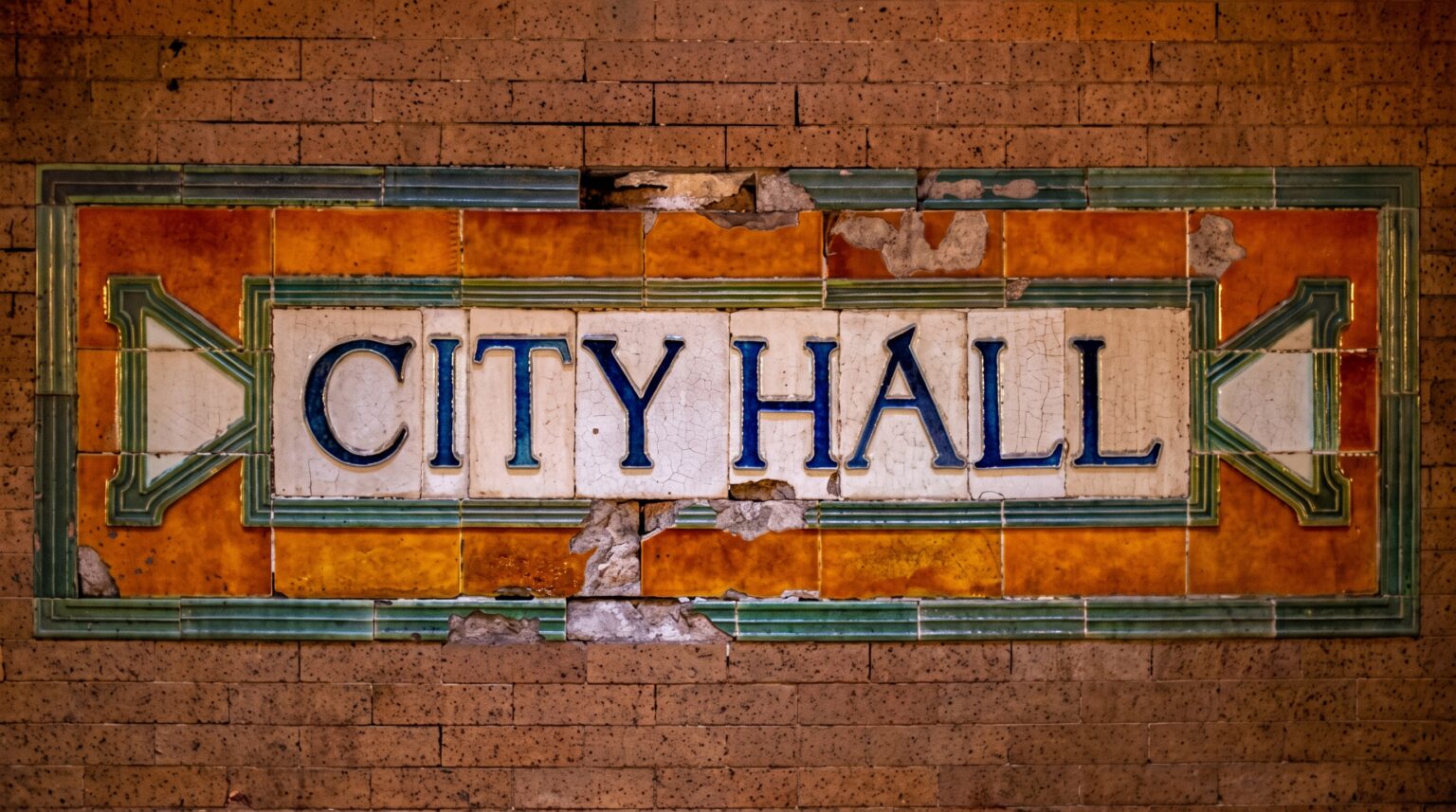 Close-up of a weathered mosaic tile sign spelling "CITY HALL" in blue letters against a white background, framed by cracked and missing orange and green border tiles that expose the raw brick underneath