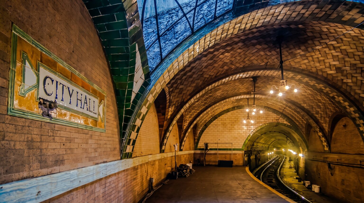A dimly lit, curving abandoned subway platform with a tiled wall featuring a mosaic sign reading "CITY HALL" in blue. The ceiling features continuous arches of herringbone-patterned brick, supported by green-tiled pillars and illuminated by vintage hanging chandeliers