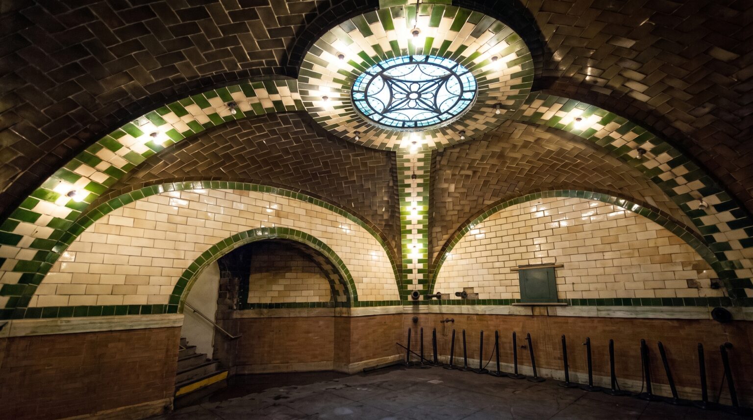 An underground chamber featuring intricate Romanesque Revival architecture, with vaulted ceilings made of beige tiles bordered by bright green tiles. A large, decorative circular glass skylight sits at the center of the ceiling overhead