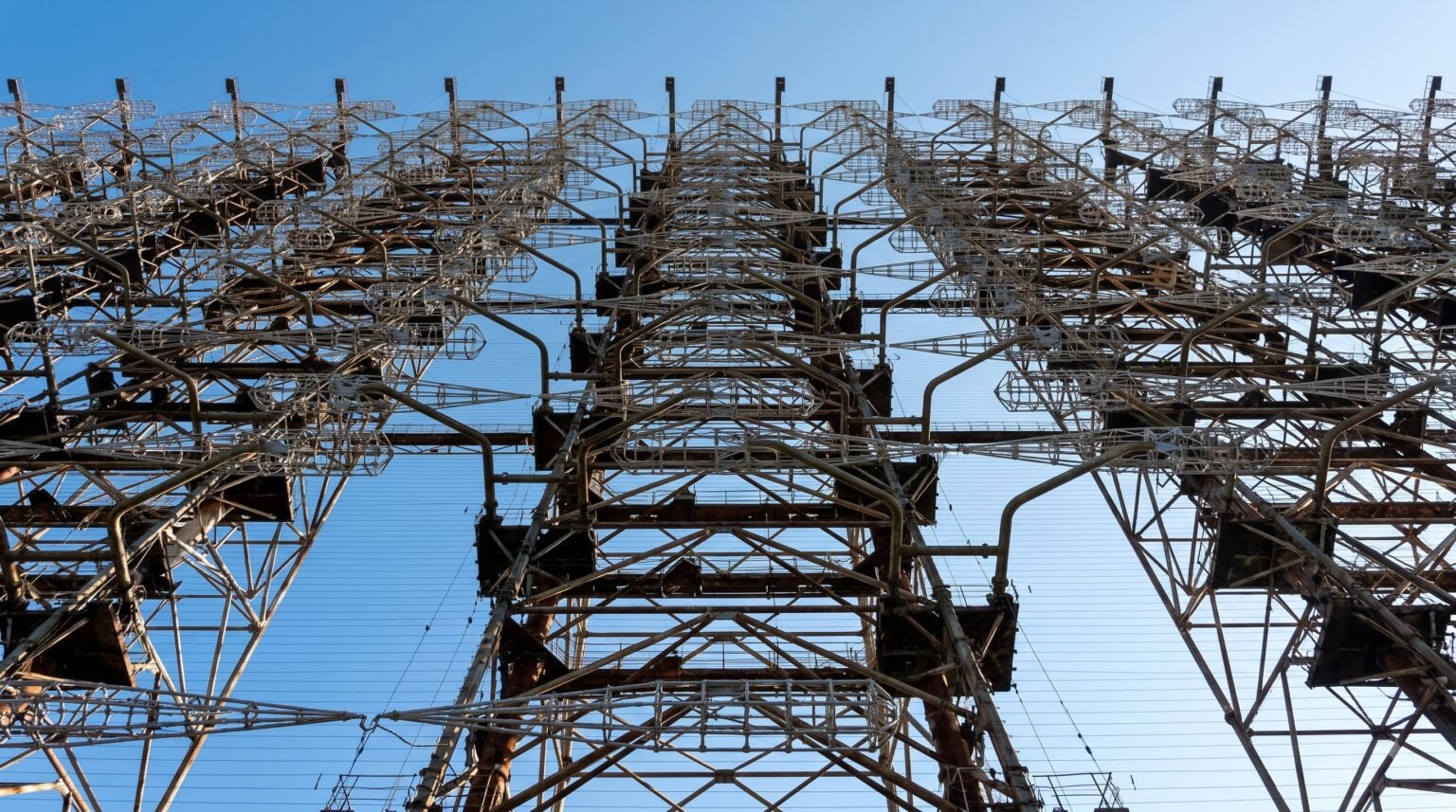 A low-angle shot looking directly up at the towering, complex steel grid and horizontal wire cages of the Duga radar against a clear blue sky