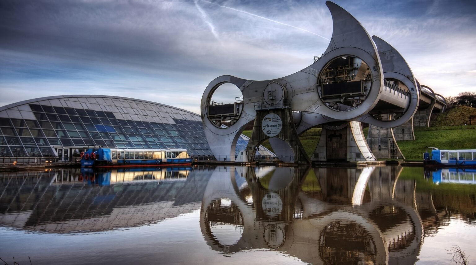 A modern rotating boat lift and adjacent glass visitor centre are perfectly reflected in the calm waters of the canal basin under a dramatic cloudy sky