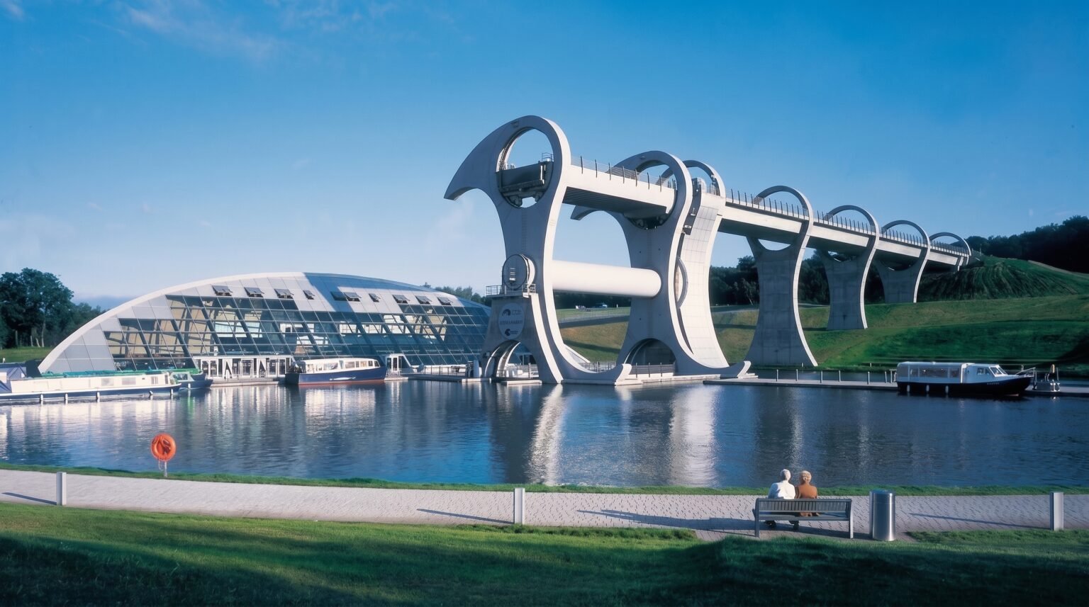 A large modern rotating boat lift stands beside a curved glass building next to a calm canal basin. Two people sit on a bench in the foreground looking at the water. Falkirk Wheel