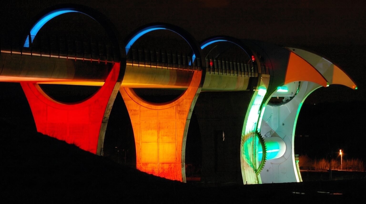 Nighttime view of a large aqueduct and boat lift structure dramatically illuminated in bright neon colors of red, orange, blue, and green