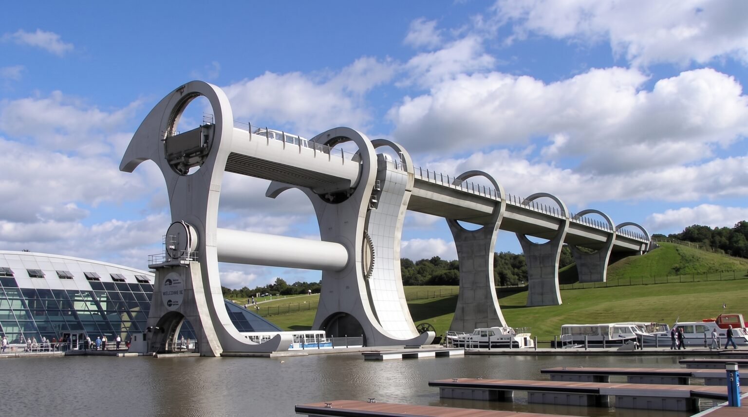 A daytime side-profile view of a massive steel rotating boat lift connecting two different elevations of a canal, with boats docked in the water below.