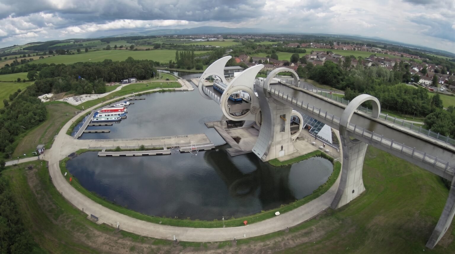 Wide aerial photograph of a rotating boat lift and its large, circular water basin, surrounded by green lawns, pathways, and trees