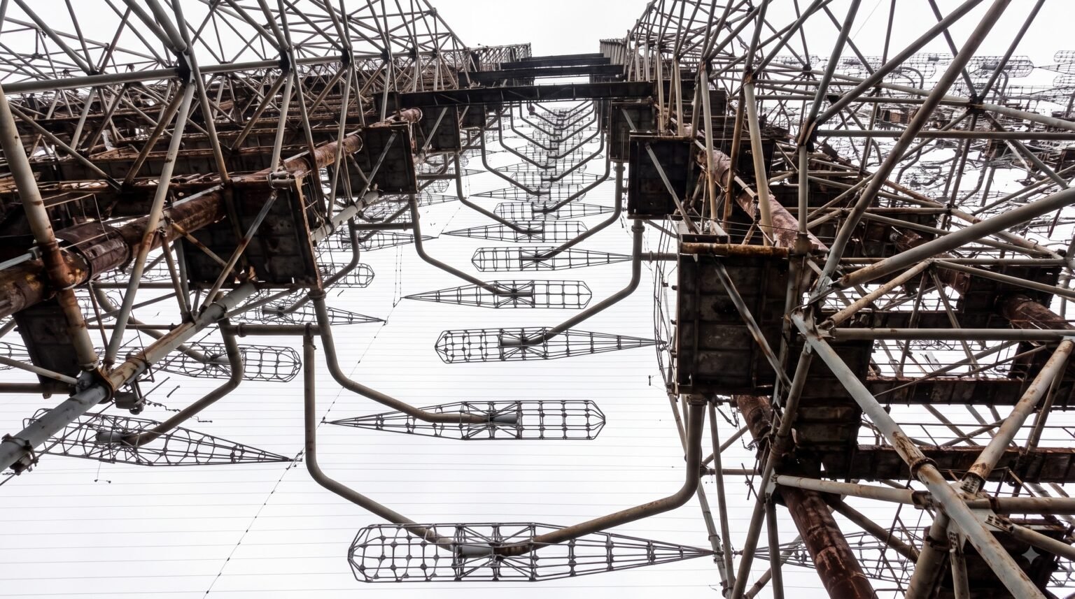 An upward-facing shot highlighting the repetitive geometric patterns of protruding cylindrical wire antennas and heavy steel scaffolding of the abandoned radar against a bright white sky
