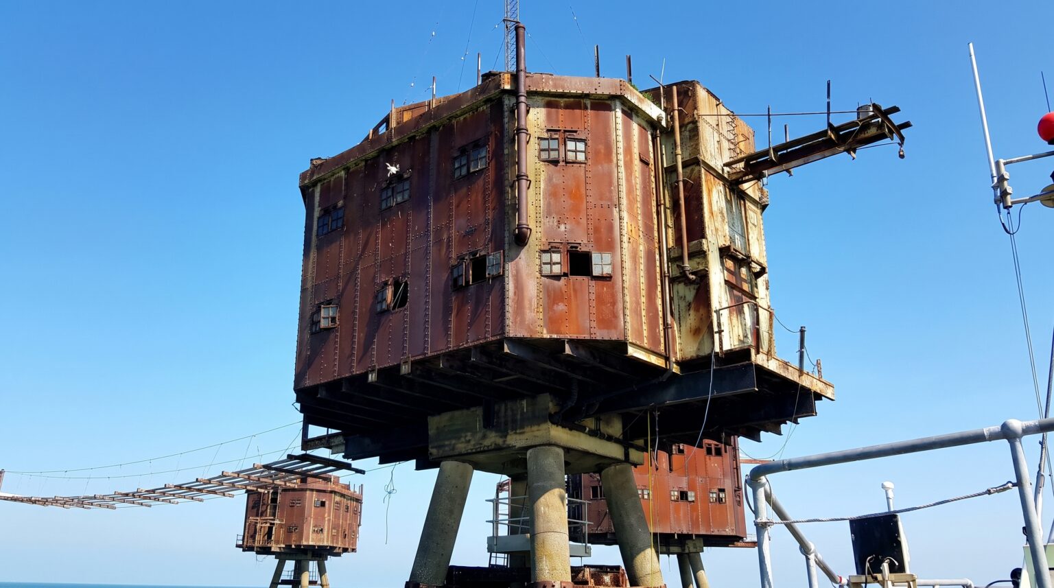 A low-angle, close-up view of a towering, deeply rusted metal sea fort resting on four sturdy concrete pillars in the ocean against a bright blue sky. A delicate, suspended wire bridge connects it to a second, partially visible fort on the left.