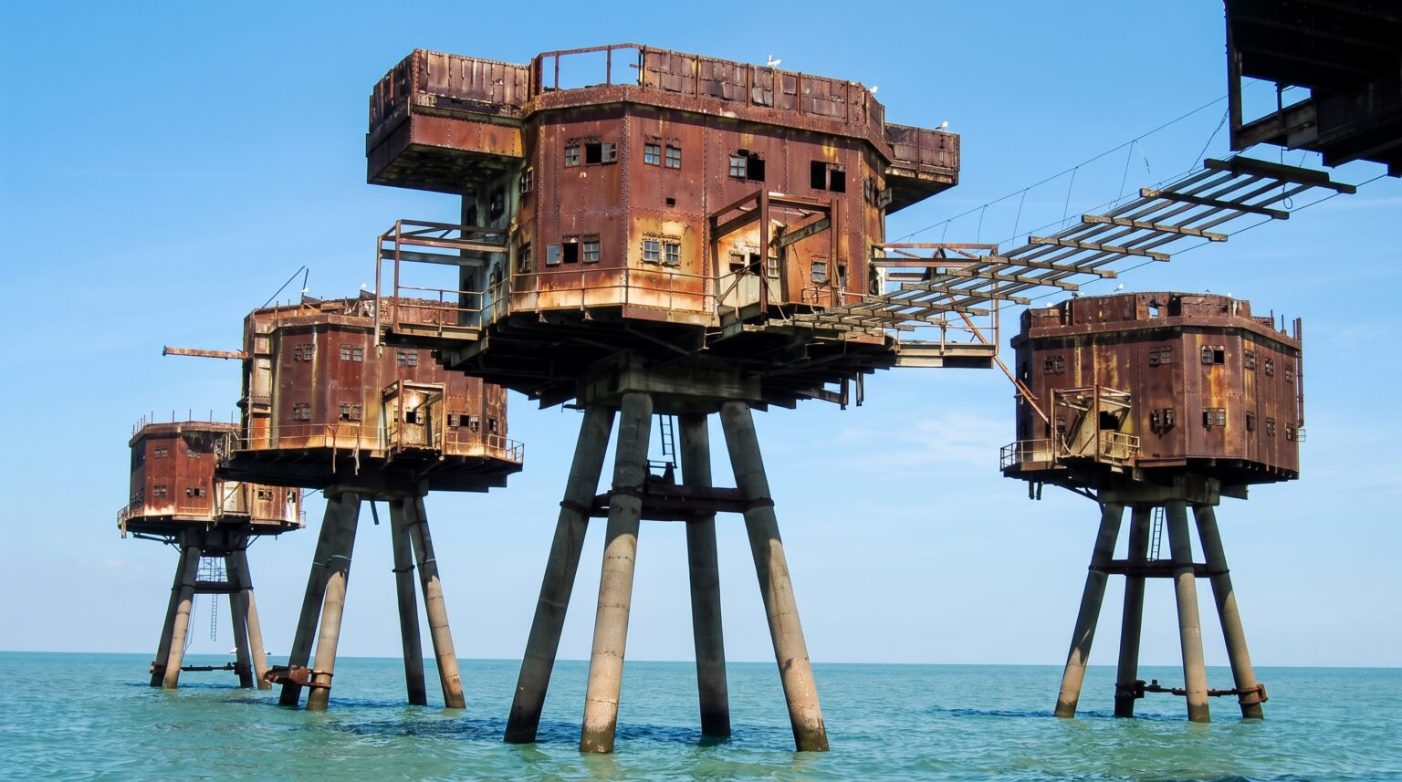 Three heavily weathered, multi-story metal sea forts standing on thick concrete stilts in the middle of a calm ocean under a clear blue sky. The boxy rusted structures feature small square windows and are connected by narrow, skeletal metal walkways.