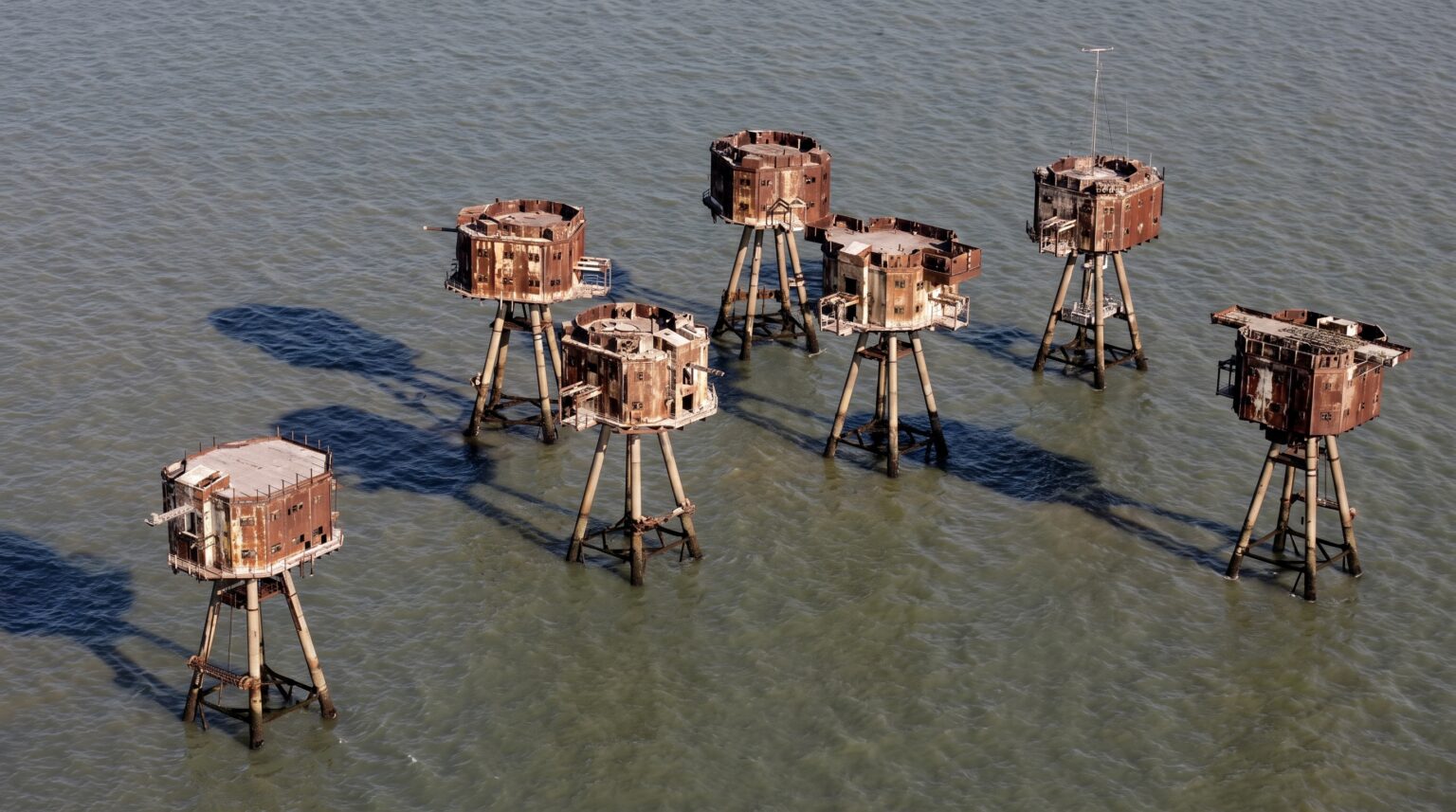 An aerial view looking down at a cluster of seven isolated, rusted sea forts standing in the ocean. The box-shaped metal structures sit atop concrete legs and cast long, distinct dark shadows across the rippling water below.