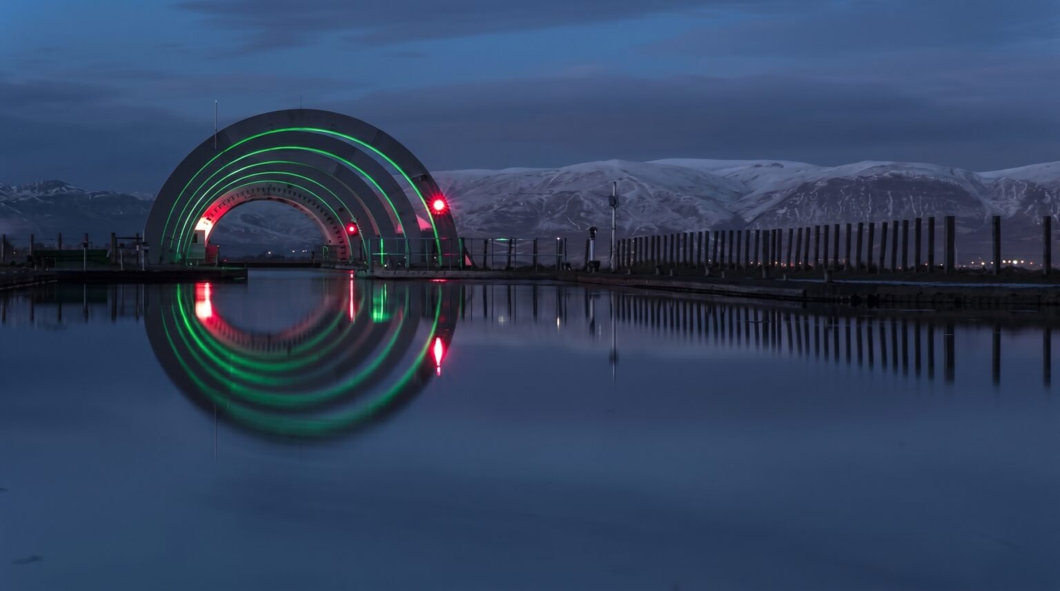 The Falkirk Wheel aqueduct illuminated with green and red neon lights at dusk, reflecting flawlessly on the calm water, with snow-capped mountains behind it