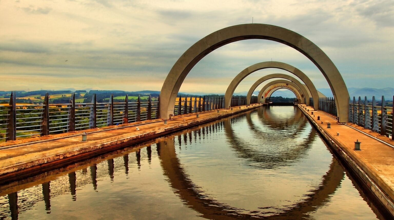 A low-angle view looking through a series of concrete arches over an aqueduct canal, with the sky and structure reflecting perfectly in the still water