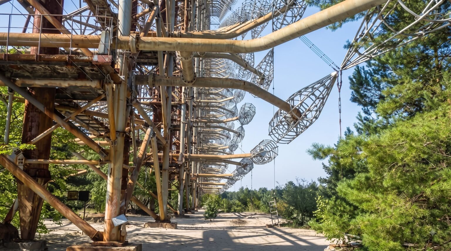 A ground-level view along the base of the massive Duga radar, showing thick rusted pipes extending outward to hold large wire cages, with trees growing near the concrete foundation