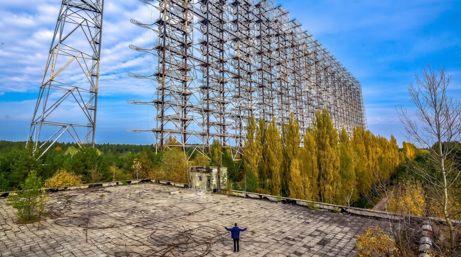 A person stands with arms wide open on a debris-covered concrete rooftop, gazing at the enormous, imposing wall of the Duga radar array surrounded by autumn trees
