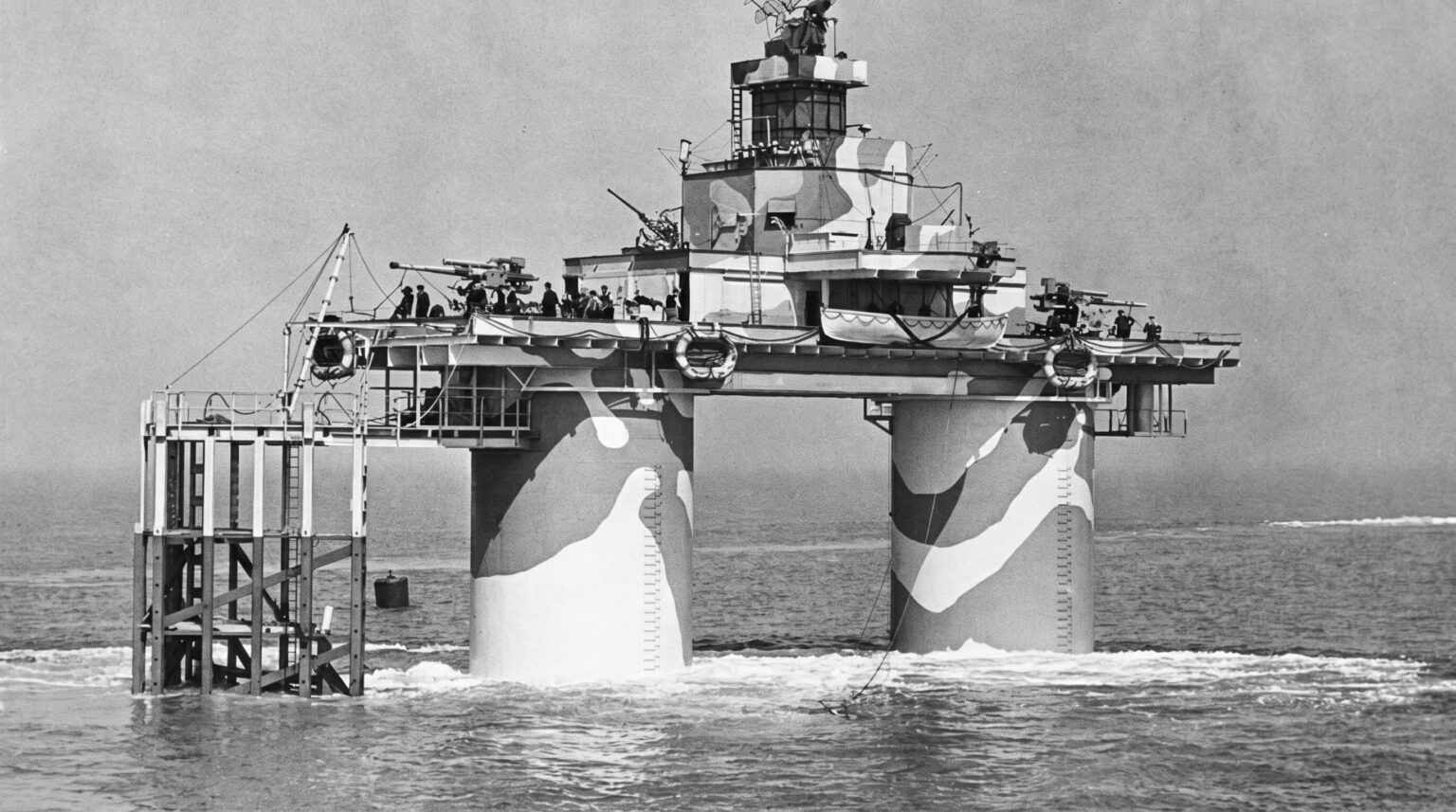 A black and white historical photograph of a military sea fort resting on two thick cylindrical pillars painted with dazzle camouflage. The upper platform is manned by naval personnel and equipped with anti-aircraft guns.