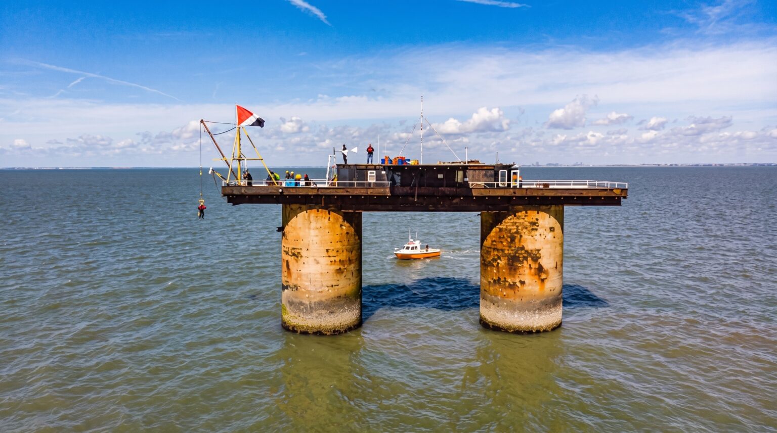 A modern color photograph of a heavily rusted offshore sea fort resting on two thick concrete pillars in the ocean. A small orange boat sails between the pillars, while people stand on the platform above, one of whom is being hoisted by a small crane next to a flying flag.