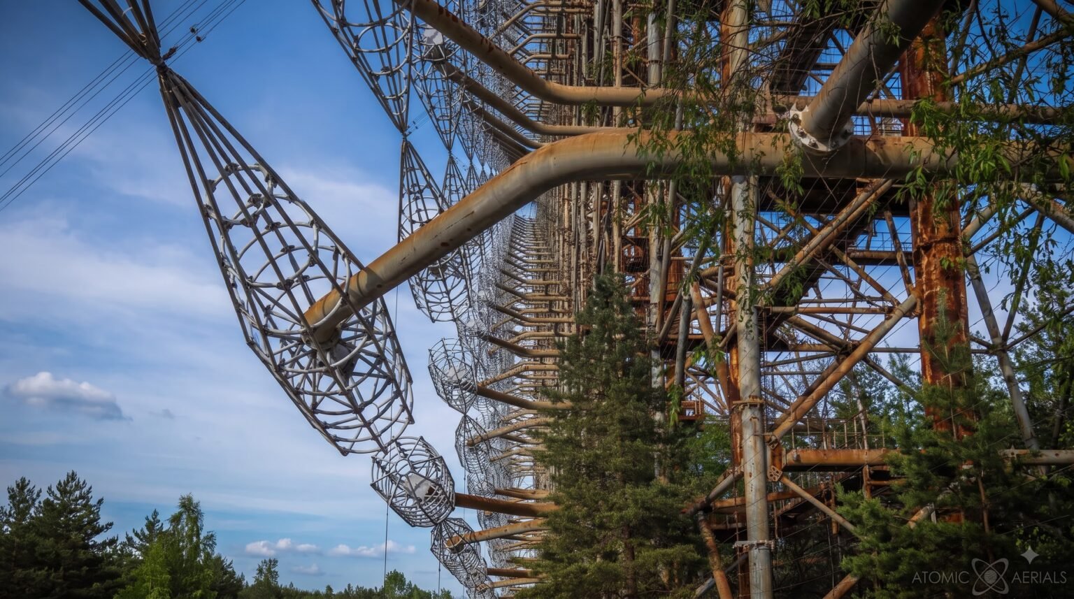 A close-up, angled view of the giant rusted arms and wire antenna cages of the Duga radar, with green pine trees growing closely amidst the decaying structure
