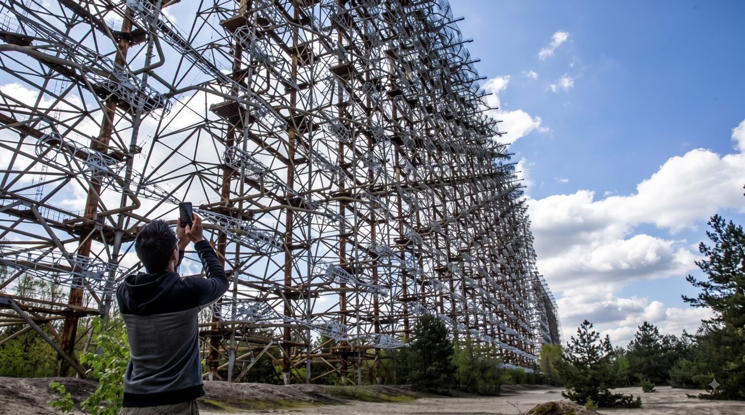 A person seen from behind takes a photo with their smartphone of the colossal, rusted steel lattice structure of the abandoned Duga radar antenna under a blue, cloudy sky