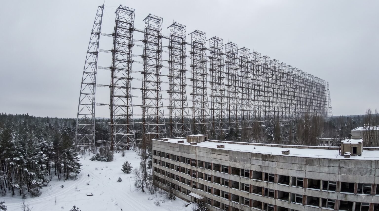 An aerial winter view of the colossal, abandoned Duga steel lattice radar array towering over a snow-covered pine forest. In the foreground sits a dilapidated, multi-story concrete building with empty window frames and a snowy roof under an overcast sky