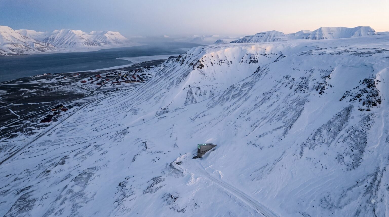 An aerial drone shot of the snow-covered Platåberget mountain in Svalbard, showing the Global Seed Vault entrance protruding from the hillside, with an icy fjord and a small town visible in the distance