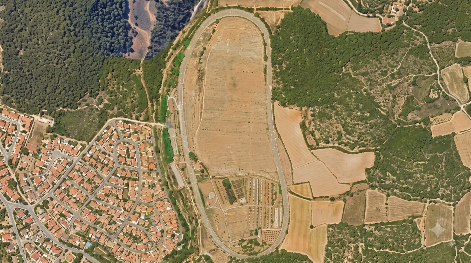 Aerial satellite view of an oval-shaped race track surrounding dry agricultural fields, situated between a dense housing development and forested hills